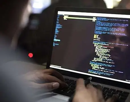 A person typing on a laptop, displaying code in a programming environment. The screen shows colorful text and lines of code in a dark theme, with a faint reflection of the individual.